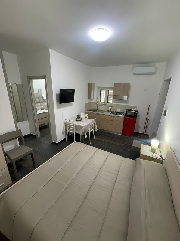 A compact hotel room with a bed, a dining table with chairs, a kitchenette, a TV mounted on the wall, and a red door.