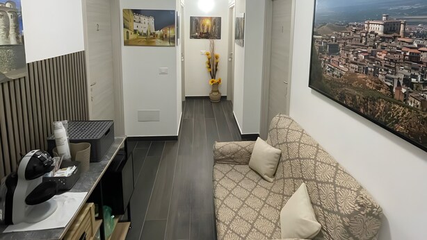 Un salon moderne avec un canapé, un bureau équipé d’une imprimante, un grand tableau représentant un paysage urbain, et un couloir menant à une autre pièce.