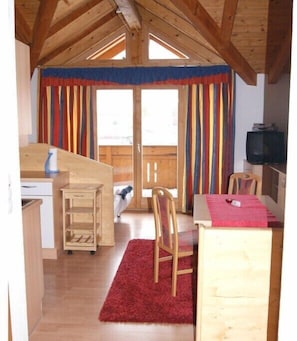 Ein kleiner, gemütlicher Raum mit Holzbalkendecke, einem Schreibtisch mit Stuhl, einem Fernseher und einem Fenster mit bunten Vorhängen.