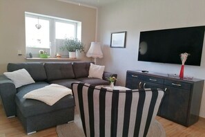 A modern living room with a sectional sofa, a striped chair, a flat-screen TV, a dark cabinet, and a window with plants. - Holiday home with 1 bedroom (Plau am See)