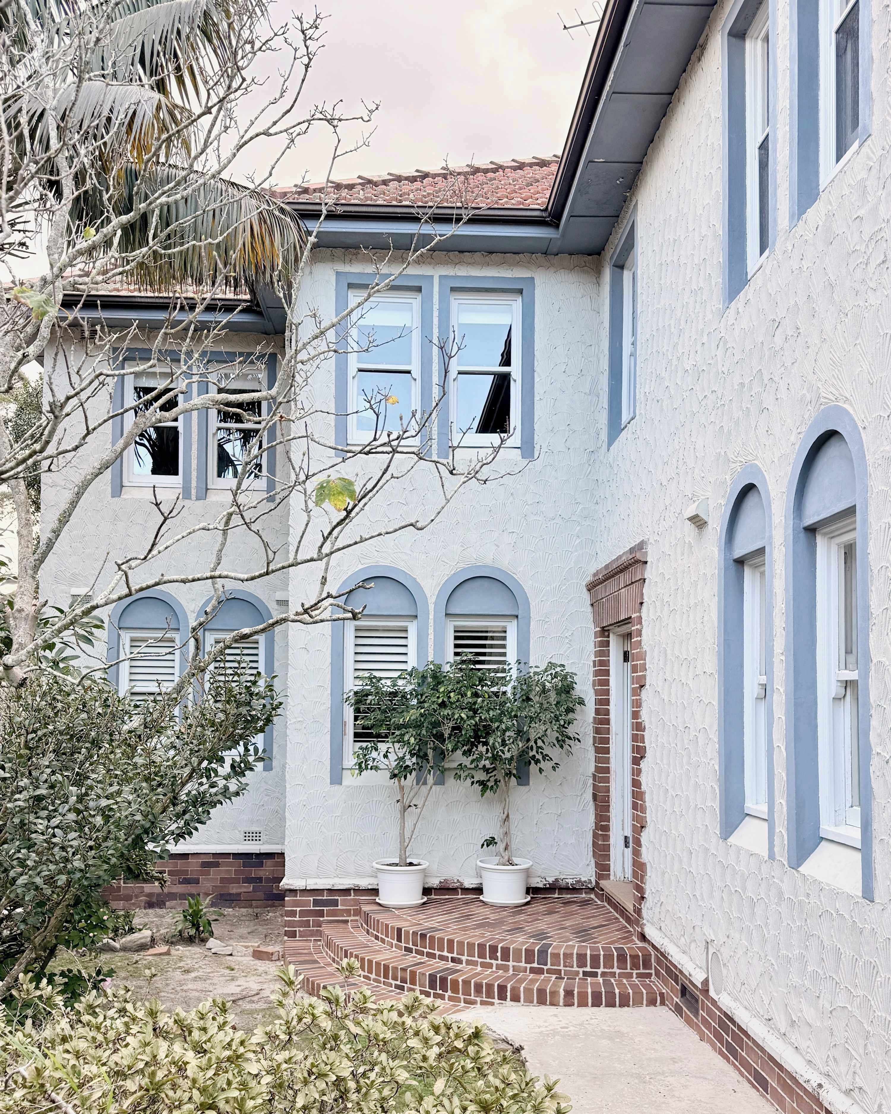 Una casa de dos pisos con fachada de ladrillo, varias ventanas y un pequeño jardín con plantas en macetas.
