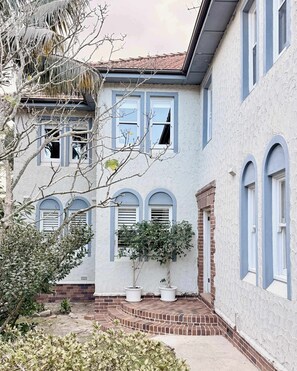 Una casa de dos pisos con fachada de ladrillo, varias ventanas y un pequeño jardín con plantas en macetas.