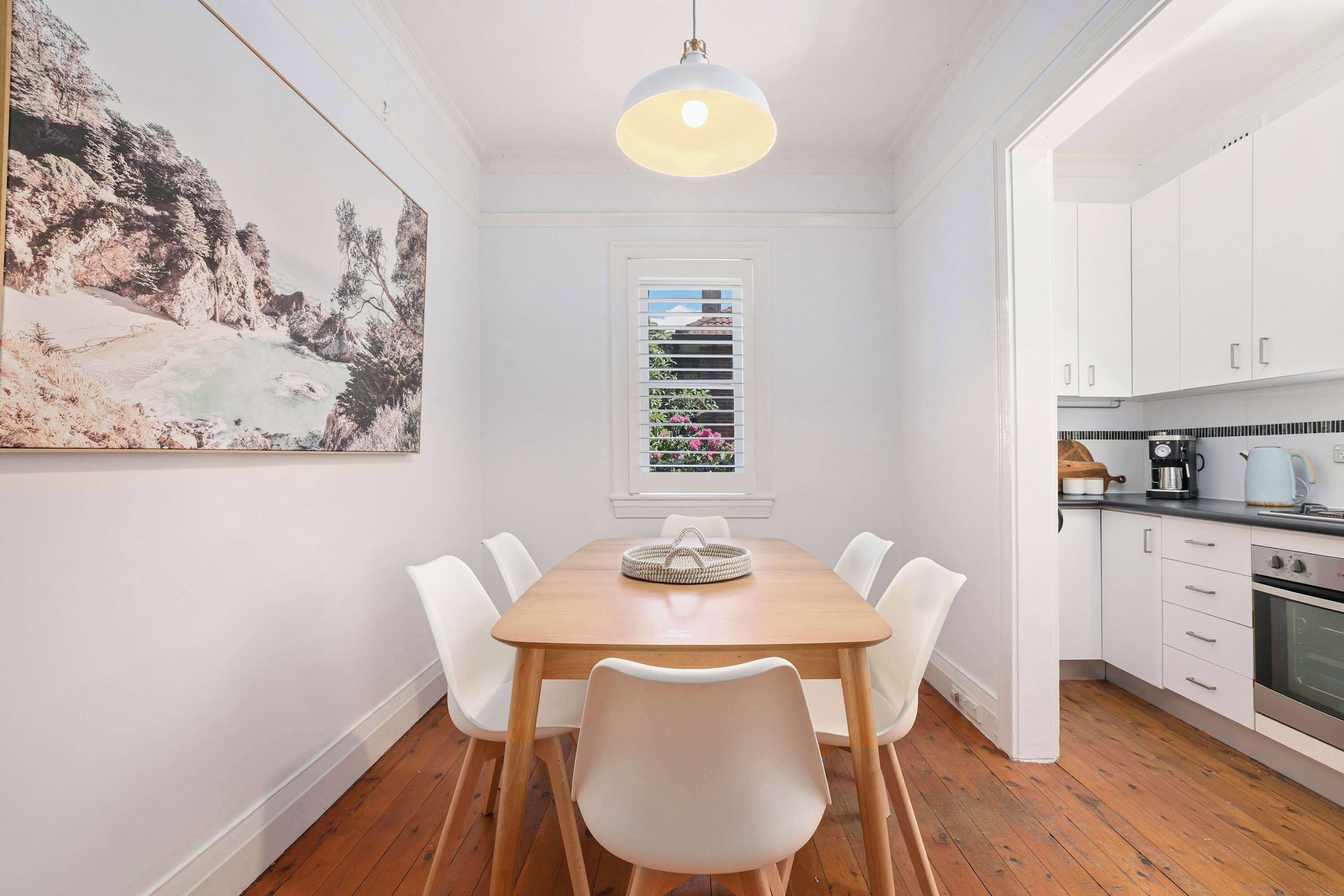 Een eetgedeelte met een houten tafel, witte stoelen, een hanglamp, een raam met zonwering en een keuken met witte kasten en apparatuur.