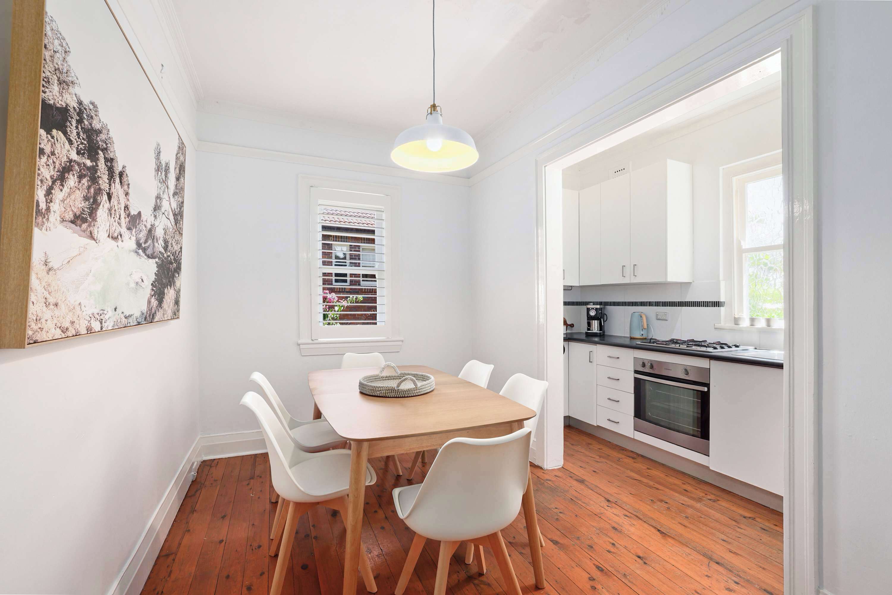 Un comedor con mesa de madera y sillas blancas, una lámpara colgante, una ventana con persianas y una cocina con armarios blancos y una estufa.