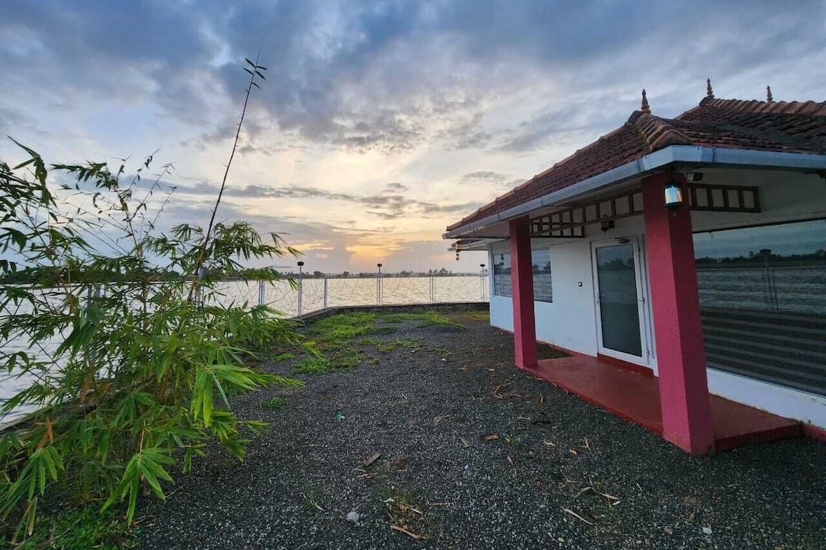 Un pequeño edificio de paredes blancas y techo rojo, ubicado cerca de una masa de agua con una valla al fondo.