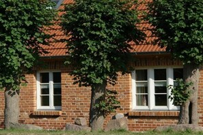 Ein Backsteinhaus mit zwei Fenstern in weißen Rahmen, einem roten Ziegeldach und umliegenden Bäumen.