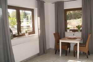 Ein Zimmer mit Esstisch, Stühlen, einem Fenster mit Vorhängen, einem Heizkörper und einem kleinen Regal mit Pflanzen.