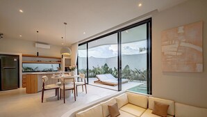 A modern interior with a kitchen, dining area, and a seating area with a view of a pool and mountains. - IVY Villa 2 (Pererenan)