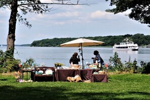 Un marché en plein air au bord du lac, avec des tables et des parasols, un bateau en arrière-plan et un arbre au premier plan.
