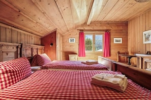 A wooden cabin room with two beds, a window with red curtains, and a small table.