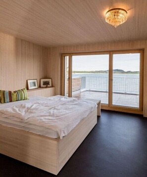 Ein Schlafzimmer mit einem großen Bett, einer Holzbalkendecke, einer Kristallleuchter und Blick auf ein Gewässer durch große Schiebetüren.