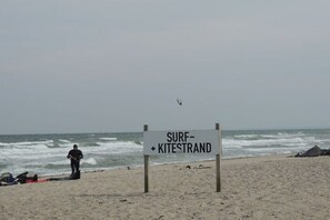 Ein Strand mit einem Schild, auf dem 'SURF+KITESTRAND' steht, eine Person am Wasser und ein Drachen am Himmel.