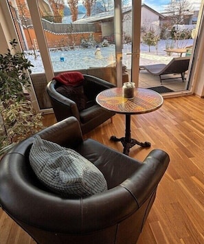A cozy room with a leather chair, a small round table, and a view of a snowy outdoor area through a large window. - Holiday apartment WH (Garmisch-Partenkirchen)