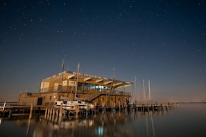 Un edificio frente al agua con un muelle techado, barcos amarrados en primer plano y un cielo nocturno estrellado.