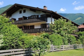Eine zweistöckige Chalet mit Balkon, umgeben von üppiger Vegetation und Bergen im Hintergrund.