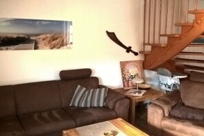 Ein Wohnzimmer mit braunem Sofa, einem kleinen Tisch mit Lampe, einem Bilderrahmen und einer Holztreppe.