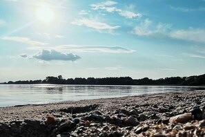 Ein Strand mit Kieselsteinen, ruhigem Wasser und einer fernen Baumlinie unter blauem Himmel.