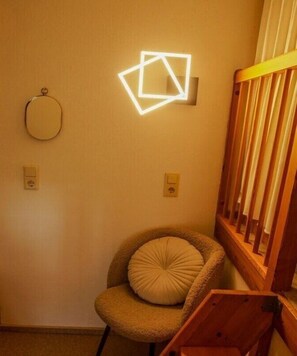Eine Ecke eines Zimmers mit einem Holzstuhl, einem Kissen und einer geometrischen Wandleuchte.