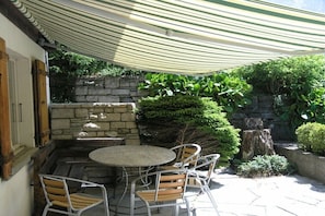 An outdoor patio with a round table and chairs, a striped awning, stone walls, and various plants.