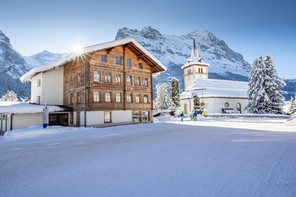 A wooden building with a snow-covered roof, a church with a steeple, and snow-covered trees in a mountainous area.