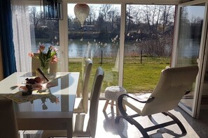 A room with a glass door leading to a garden with a pond and birds, a table with a vase of flowers, and a chair. - Holiday home with 2 bedrooms (Parey\/Elbe)