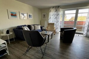 Ein Wohnzimmer mit einer Couch, Sesseln, einem Couchtisch und einem Fernseher. An den Wänden hängen Bilder in Rahmen, und ein Fenster ist mit Vorhängen versehen.