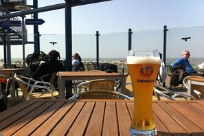 Ein Bierglas auf einem Holztisch mit Außenplätzen, mit Blick auf einen Strand und einen klaren Himmel.