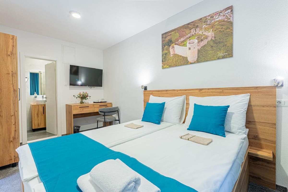 Un lit double avec une literie bleue et blanche, une tête de lit en bois, un bureau avec une chaise, une télévision et un tableau accroché au mur.
