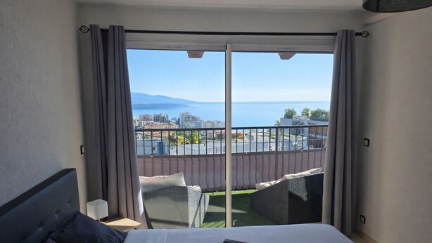 Habitación de hotel con cama, vista a la ciudad y al mar, y balcón con sillas.