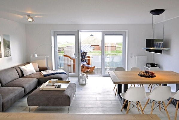 A modern living room with a sofa, coffee table, and dining area with chairs. There is a TV mounted on the wall and a large window providing a view of the outdoors. - Holiday apartment in Juist with swimming pool (Juist)
