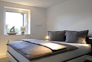 A modern bedroom with a bed, a nightstand, a window with a view, and a small table with a lamp. - Holiday apartment in Juist with swimming pool (Juist)