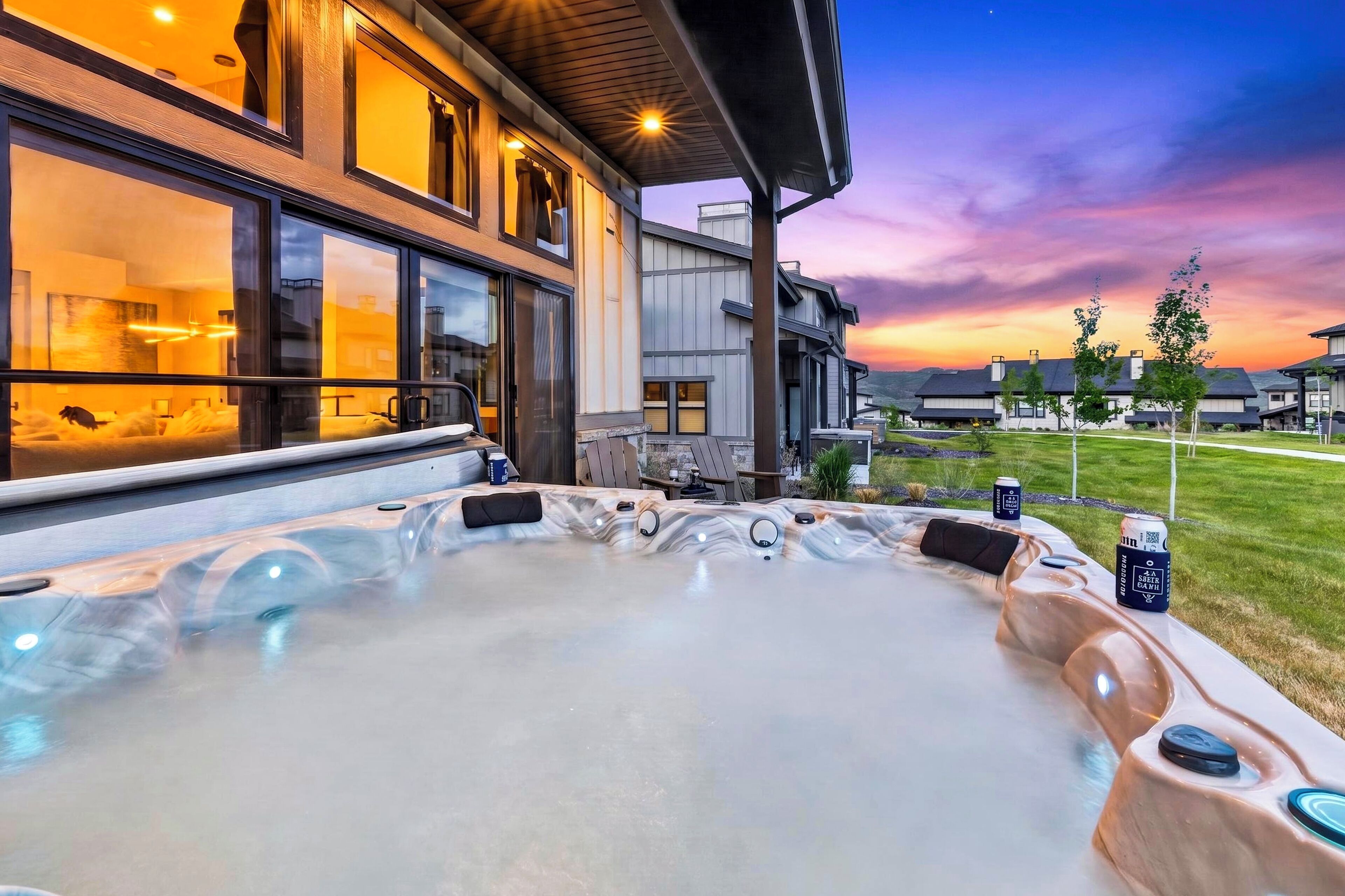 Een bubbelbad met zitplaatsen en bedieningselementen, een modern huis met grote ramen, en uitzicht op een zonsondergang.