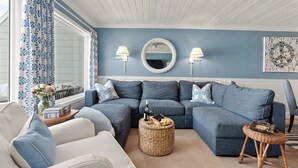 Un salon avec un canapé bleu, un fauteuil blanc, un pouf rond en osier et une table d’appoint sur laquelle se trouvent une bouteille et des en-cas.