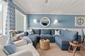 Ein Wohnzimmer mit einem blauen Sofa, einem weißen Sessel, einem runden Weidenhocker und einem Beistelltisch mit einer Flasche und Snacks.