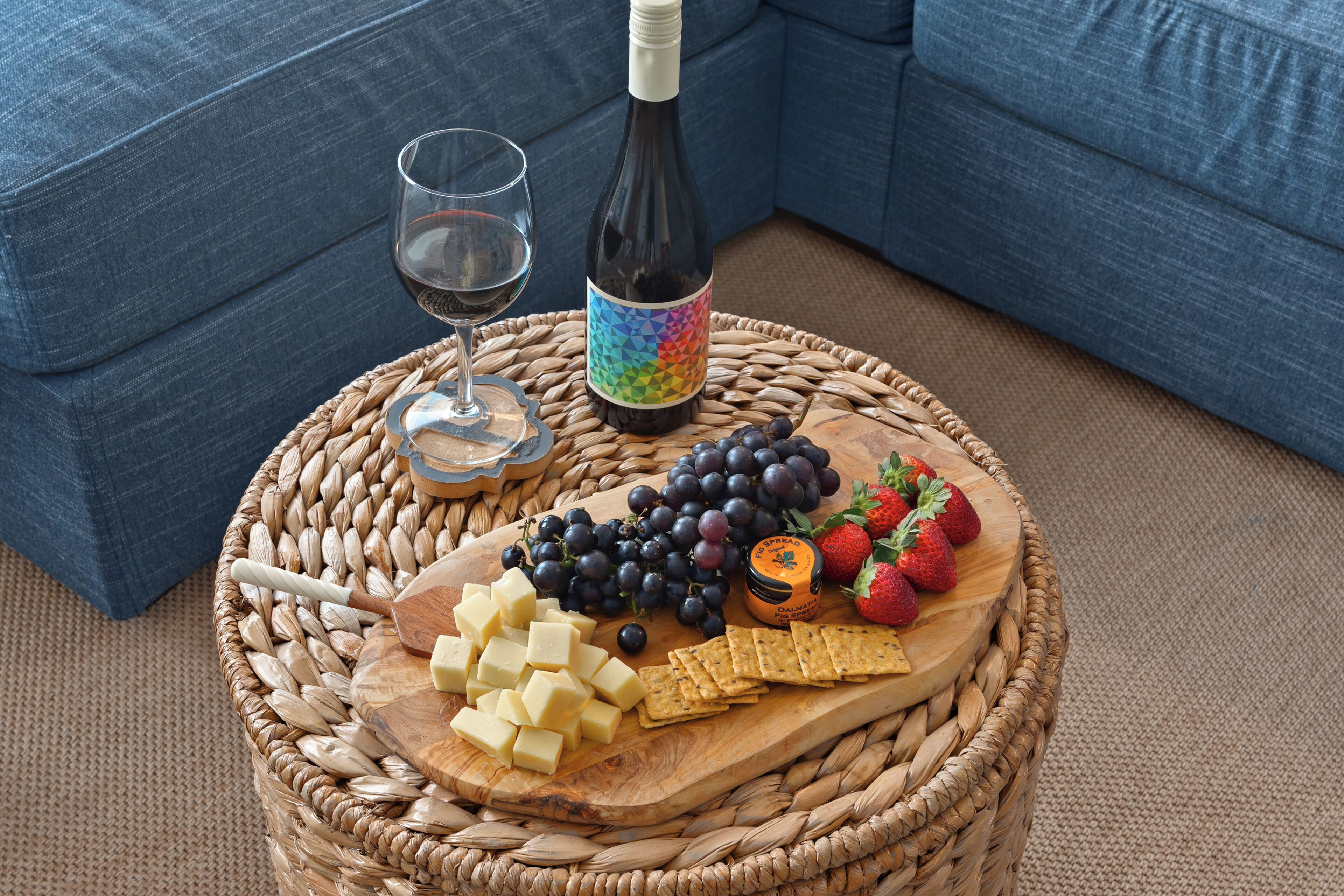 A wicker basket with a wooden board containing cheese, crackers, grapes, strawberries, and a bottle of wine on a woven mat.