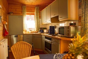 Eine kompakte Küche mit Kühlschrank, Mikrowelle, Backofen und einem Essbereich mit einem Korbstuhl.