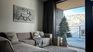 Een moderne woonkamer met een grijze bank, een open haard en een groot raam met uitzicht op een besneeerd landschap.