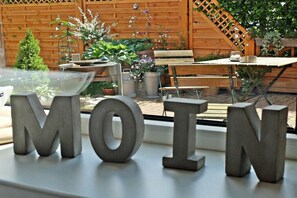 Eine Terrasse mit Holzzaun, Kübelpflanzen, einer Bank und großen Metallbuchstaben, die 'MOIN' auf einem Tisch bilden.