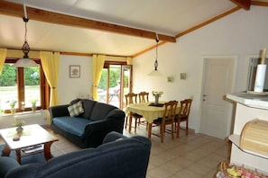 A living room with a sofa, a dining table with chairs, a kitchen area, and a window with curtains. - Holiday home in De Haan with swimming pool (De Haan)
