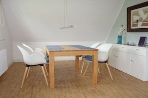 A dining area with a wooden table and white chairs, a white cabinet, and a framed picture on the wall. - "Seagull under thatch" (Nebel)