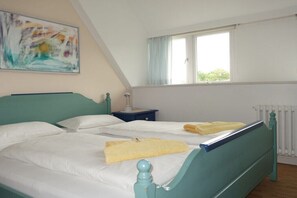 A bedroom with two beds, a window with curtains, a radiator, and a painting on the wall. - "Seagull under thatch" (Nebel)