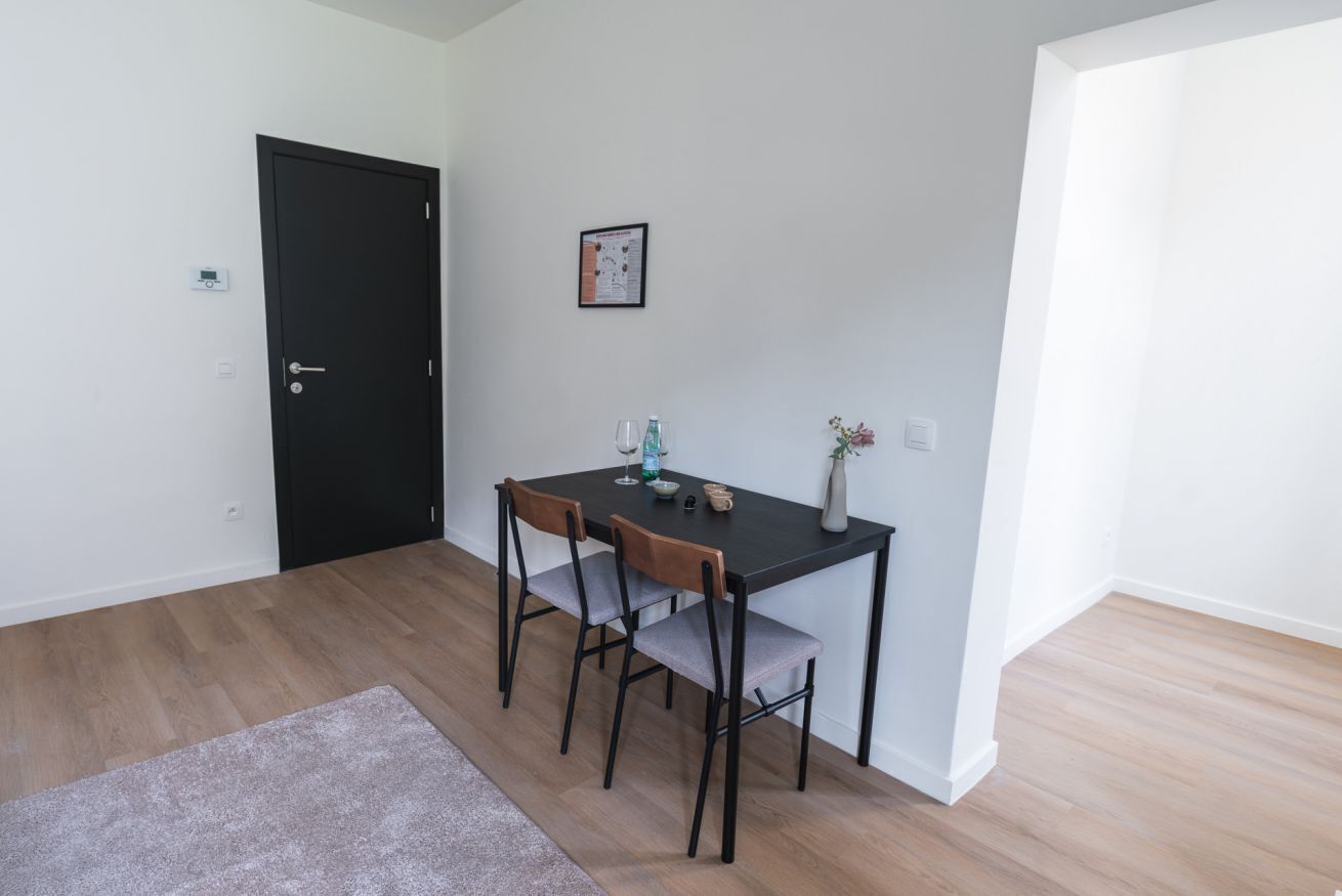 Une pièce avec une porte noire, un tableau encadré au mur, une table avec deux chaises et un petit vase.