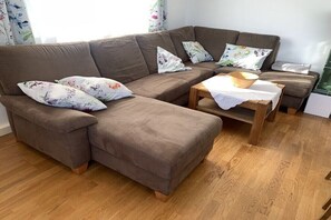 Un canapé d’angle marron agrémenté de coussins à motifs floraux, une table basse en bois avec un bol, et un plancher en bois.