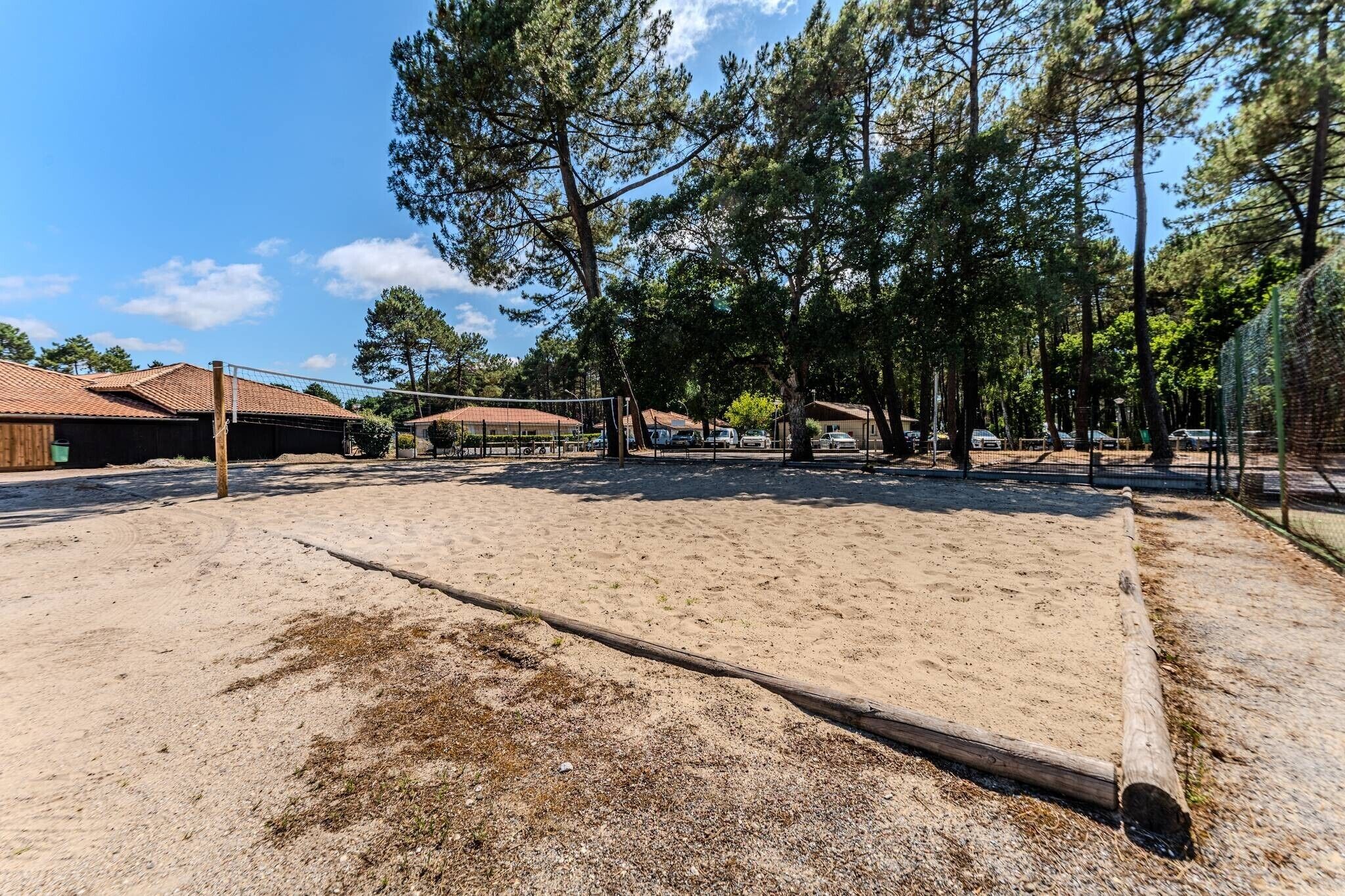 A sandy outdoor area with a volleyball net, surrounded by trees and buildings with red-tiled roofs.