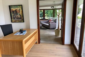 Ein Holztisch mit einem Stuhl, ein gerahmtes Kunstwerk an der Wand und Blick auf einen Balkon mit einer Couch und einem Topfpflanze.