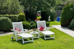 Two white lounge chairs with red cushions, a small white table, and a potted plant in a well-manicured garden with hedges and a statue.