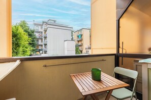 Ein Balkon mit einem Holztisch und einem grünen Glas, mit Blick auf eine Stadtlandschaft aus Gebäuden und Grünflächen.
