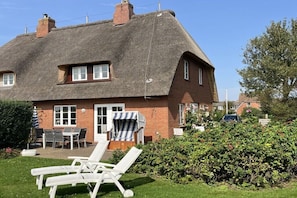 Ein Haus mit einem Reetdach, einer Terrasse mit Tisch und Stühlen und einer Hecke im Vordergrund.