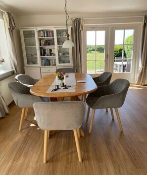 Ein Essbereich mit einem runden Holztisch, grau gepolsterten Stühlen, einer weißen Kommode mit Glastüren und Blick ins Grüne.