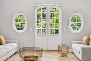 Ein Zimmer mit einer weißen Tür, zwei runden Fenstern, einem weißen Sofa und einem runden Tisch mit goldener Basis.